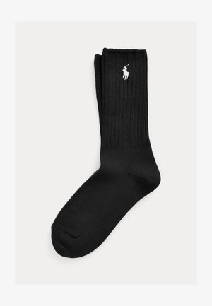 Par črnih rebrastih nogavic s črto in belega vezenega logotipa igralca polo blizu zgornjega roba manšete.