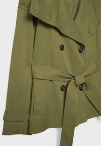 Trench-coat vert olive avec boutons à double boutonnage, larges revers, taille ceinturée et détails à brides sur les poignets.