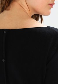 Schwarzer Pullover mit weitem Ausschnitt, der ein strukturiertes Strickmuster und kleine Knöpfe am Rücken hat. Das Material wirkt weich und leicht.