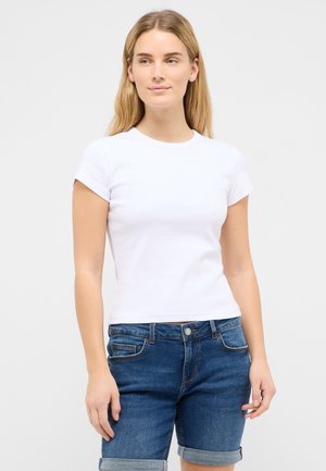 Frau mit langen blonden Haaren, die ein einfaches weißes T-Shirt mit kurzen Ärmeln und hochgekrempelte blaue Jeansshorts vor einem einfarbigen Hintergrund trägt.