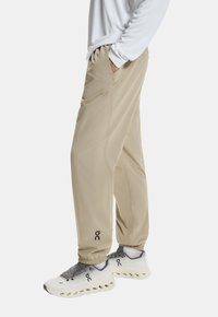 Beżowe spodnie sportowe z elastycznymi mankietami i bocznymi kieszeniami, wykonane z lekkiego materiału. Zawierają logo na dolnej części nogawki. W połączeniu z szarymi butami sportowymi.