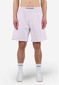 Gaiši rozā sporta šorti ar elastīgu jostu. Ir sānu kabatas un logotips pie apakšējās malas, valkāti kopā ar baltām sporta kurpēm un zeķēm.