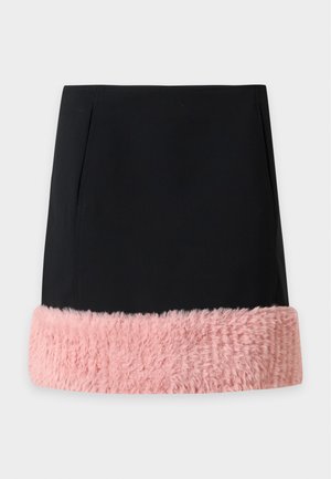 Kvinde iført en pink t-shirt med grafik, sort mininederdel med pinke lodne detaljer og sorte sandaletter med hæl, stående mod en ensfarvet baggrund.