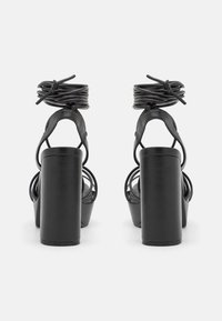 Sandalias de tacón alto negras con un tacón grueso, tiras delgadas alrededor del tobillo y detalles envueltos en la parte posterior. Textura de cuero suave y diseño minimalista.