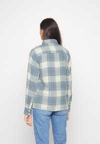 Koszula w kratę w niebieskich i jasnopylnych odcieniach, wykonana z teksturowanej tkaniny. Posiada standardowy kołnierz oraz mankiety zapinane na guziki. Widoczne szwy i wzory.
