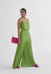 Groene geplooide jumpsuit met V-hals, gecombineerd met een roze textuur clutch en roze gestreepte sandalen met hakken, voorzien van een kettinghengsel.
