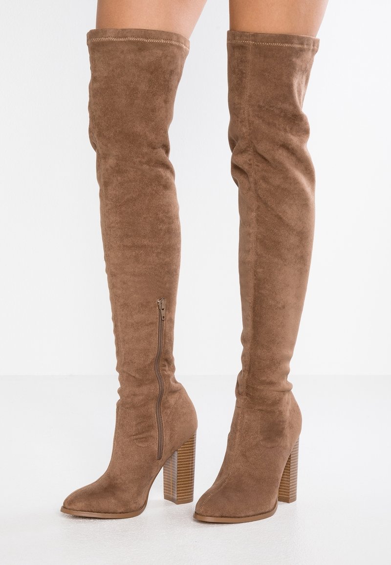 Stivali alti in suede marrone con tacco, cerniera laterale e tacco a blocco su uno sfondo bianco.