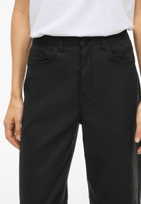 Juodos kelnės iš lygaus audinio, vidutinio aukščio liemeniu, su priekinėmis kišenėmis ir subtiliomis siuvimo detalėmis. Klasikinis dizainas su aptempiamu stiliumi.