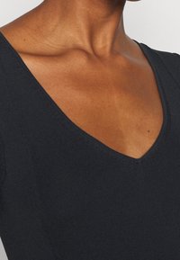 Top tricoté noir avec un décolleté en V profond, présentant une texture côtelée et une finition lisse sur les bords. Accent sur le tissu et les détails du décolleté.