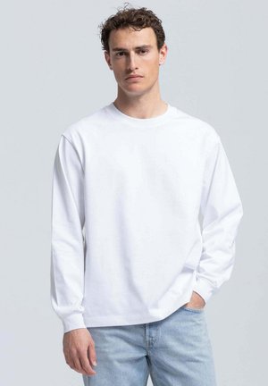Jonge man met krullend haar, die een eenvoudig wit shirt met lange mouwen en lichtblauwe spijkerbroek draagt, staand met één hand in zijn zak.