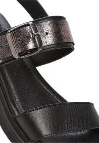 Sandale en cuir noir avec une large bride d’orteil et une bride de cheville métallique brillante fixée par une boucle métallique rectangulaire.
