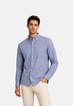 Ung mand står med den ene hånd i lommen iført en lyseblå skjorte med knapper og beige bukser mod en ensfarvet baggrund.