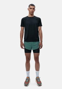 Juoda trumpa rankovė margo, žalia ir juoda šortai, pilki sportiniai batai su oranžiniais akcentais, balti sportiniai kojinės, vienspalvis fonas.