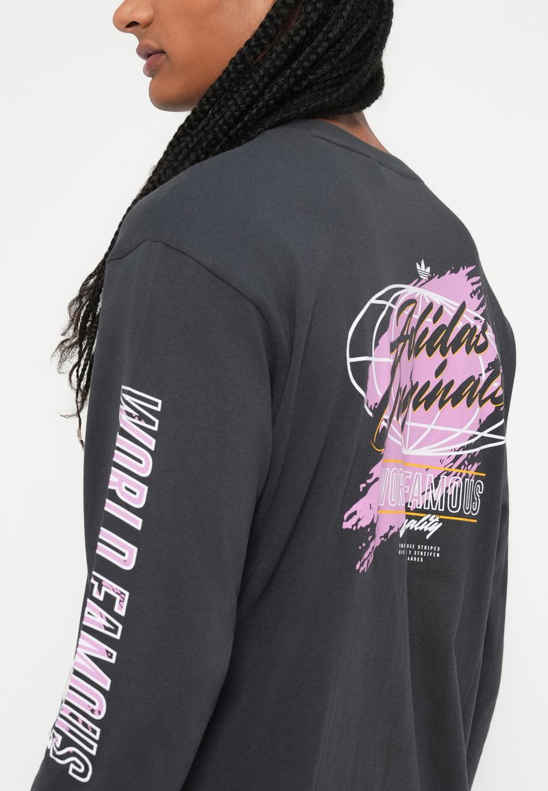Frau mit geflochtenen Haaren, die ein schwarzes Langarmshirt mit pinkem und weißem "World Famous"-Text auf dem Ärmel und einem grafischen Design auf dem Rücken trägt.