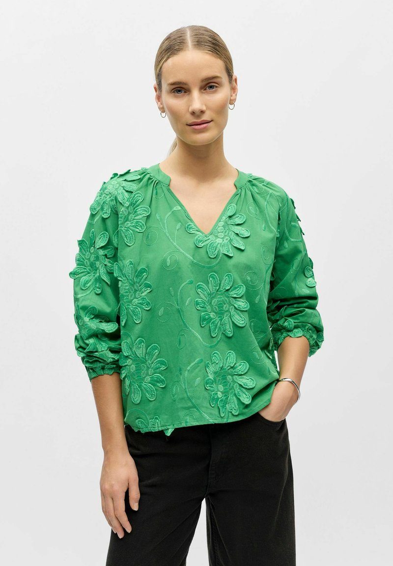 Femme aux cheveux blonds portant un chemisier vert brodé de fleurs avec des manches retroussées et un pantalon noir, se tenant avec une main dans la poche.