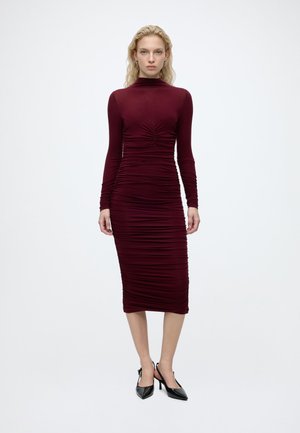 Femme aux cheveux blonds portant une robe midi ajustée, à manches longues, froncée bordeaux et des chaussures à talons hauts slingback noires, debout sur un fond blanc.