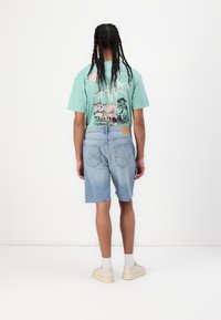 Tricou grafic turcoaz cu un design roz, asortat cu pantaloni scurți din denim de culoare albastru deschis și sneakers albi cu talpă bej.