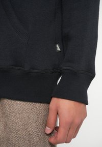 Felpa nera con texture, dotata di tasca a marsupio, polsini e orlo a coste. Presenta una piccola etichetta del marchio sul lato. Sono visibili pantaloni marroni con fantasia.