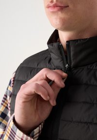 Colete acolchoado preto com colarinho alto, apresentando um fecho frontal. Uma camisa quadriculada com detalhes em vermelho, amarelo e azul é visível por baixo.