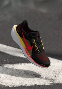 Schwarzer, roter und gelber Sneaker mit einem Netzobermaterial, strukturiertem Design und einem auffälligen roten Swoosh. Weiße und schwarze Laufsohle mit "AIR ZOOM"-Detail.