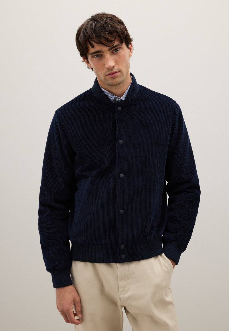 Giacca in velluto a coste blu navy con colletto rotondo, bottoni frontali e due tasche laterali. Presenta polsini a coste e una fodera interna liscia.