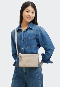 Ung kvinna i denimskjorta och jeans, leende, bär en beige axelväska med dragkedja och runt logodetalj.