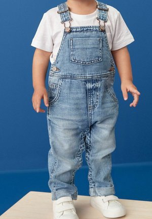 Tout-petit portant une salopette en denim bleu, un t-shirt blanc et des baskets blanches, debout sur une plateforme en bois devant un fond bleu.