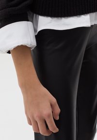 Suéter negro de canalé sobre una camisa blanca con cuello y puños de volantes, mostrando una mano descansando junto a unos pantalones de cuero sintético negros.
