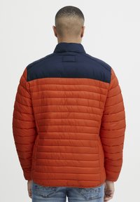 Polstret jakke med orange og navy farveblokke, der har en quiltet tekstur, en stående krave og sidelommer. Vist fra ryggen.