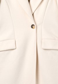 Blazer color crema chiaro con tessuto texturizzato, con chiusura a bottone singolo e due tasche piccole sul davanti.