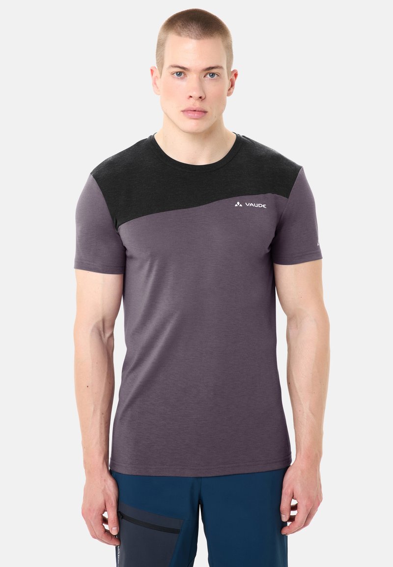 Jeune homme aux cheveux courts portant un t-shirt à manches courtes Vaude violet et noir et un pantalon bleu avec une poche zippée.