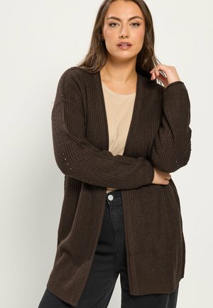 Kvinde med langt brunt hår iført en mørkebrun strikcardigan over en beige skjorte og sorte jeans, poserer med armene krydset og den ene hånd nær ansigtet.