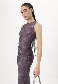 Αμάνικο, εφαρμοστό φόρεμα σε σκούρο μωβ με υφές κυματιστών σχεδίων και πλαϊνές πτυχώσεις; συνδυασμένο με μια ασημένια τσάντα.