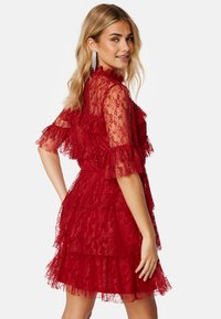 Abito rosso in pizzo con maniche corte e volants e strati. La texture è intricata, con motivi floreali e un busto aderente.