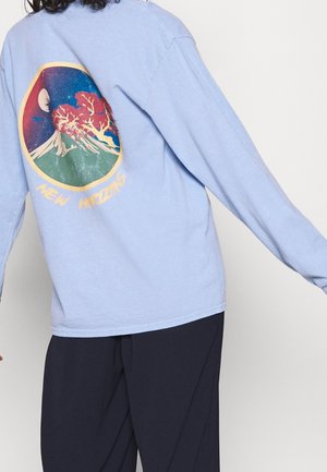 Personne portant une chemise bleu clair à manches longues avec un motif coloré de montagne et d'arbre et le texte "New Horizons" dans le dos.