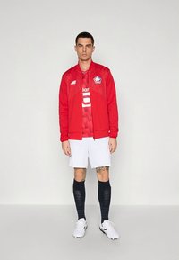 Rød atletisk jakke med hvit logo, rød drakt med nummer, hvite shorts og svarte knelange sokker; vist på en nøytral bakgrunn.