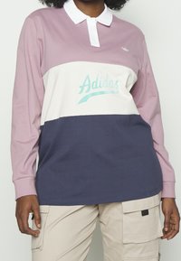 Langärmeliges Poloshirt in Lavendel, Creme und Marineblau. Verfügt über einen weißen Kragen und ein türkisfarbenes Adidas-Logo. Aus Baumwollmischgewebe.
