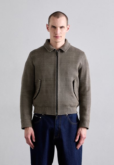 Braune gemusterte Zip-Jacke mit Kragen, zwei Seitentaschen und gerippten Bündchen, kombiniert mit dunkelblauen Jeans. Glatte Textur, strukturierte Passform.