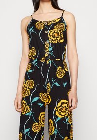 Černý overal s žlutým květinovým potiskem, tenkými ramínky, zapínáním na knoflíky vpředu a elastickým pasem. Měkká, lehká látka.