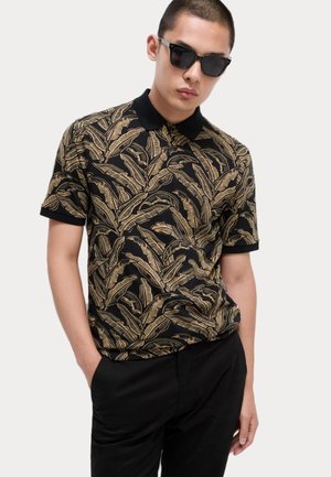 Polo negro con patrón de hojas doradas, con un cuello clásico y mangas cortas, combinado con pantalones negros. Diseño de tejido texturizado visible.