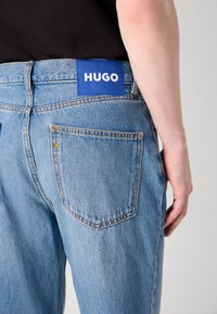 Világoskék farmernadrág egyenes szárú szabással, látható varrással, és egy kék címkével, amelyen a "HUGO" szó olvasható a hátsó derékrészen.