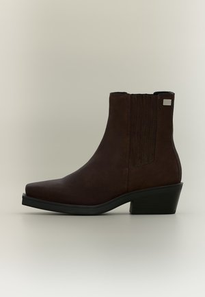Stivaletto alla caviglia in camoscio marrone scuro con tacco basso, pannelli laterali elastici e punta quadrata su sfondo grigio chiaro.