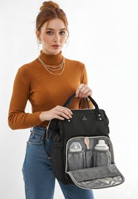 Sort multifunktionel bleetaske med quiltet tekstur, der har et frontrum med to flasker og en ske i grå holder. 