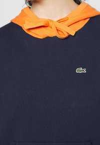 Navy sweatshirt with an orange drawstring hood, featuring a small green crocodile logo on the chest. Soft texture and relaxed fit.