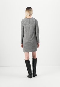 Woman with blonde hair wearing a gray long-sleeve dress and black knee-high boots, standing facing a white wall.