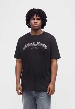 Jeune homme aux cheveux bouclés et foncés portant un t-shirt noir Jack & Jones et un jean bleu clair, debout devant un fond uni.