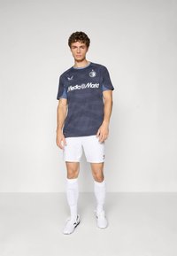 Jaunas vyras stovi vilkėdamas tamsiai mėlyną raštuotą sportinį marškinėlį su "Media Markt" logotipu, baltas šortus, kelių kojines ir futbolo batelius ant lygaus fono.