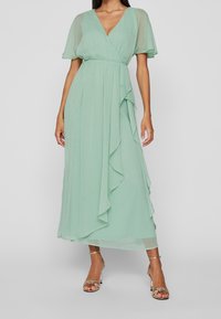 Vrouw in een mintgroene midi-jurk met doorschijnende korte mouwen, V-hals, wikkel taille, ruches en gouden enkelbandhakken tegen een grijze achtergrond.