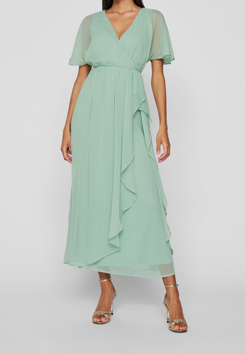 Vrouw in een mintgroene midi-jurk met doorschijnende korte mouwen, V-hals, wikkel taille, ruches en gouden enkelbandhakken tegen een grijze achtergrond.