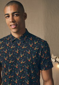 Korteærmet sort polo-shirt med farverigt hjortehovedmønster, prydet med stjerner og kristtorn, med en knapkrave og blød tekstur.